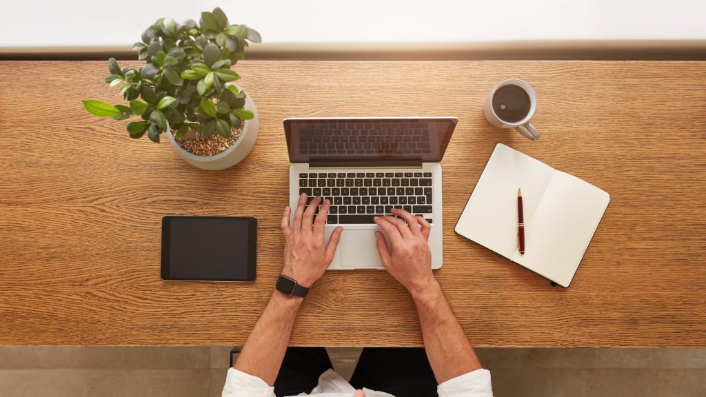 A man working on his laptop on a wooden desk next to a plant. A virtual office in Singapore encourages utilisng remote teams.