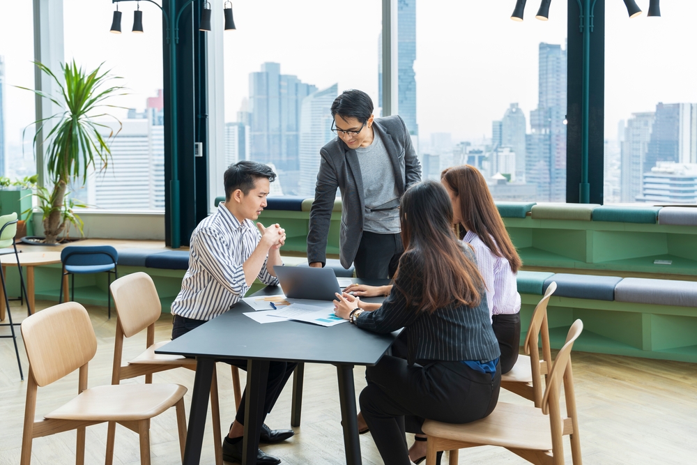 Employees collaborating over a table in a green, forest-colored coworking space. One man stands over them, operating the laptop. A virtual office solution in Singapore can't be a physical workplace for employees.