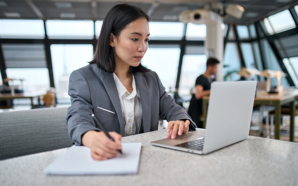 Lady in an office going through her laptop and taking notes with a pen and paper, comparing a physical office and a virtual office in Singapore