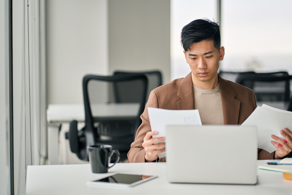 Businessman thinking, comparing two sheets of paper. He's weighing the pros and cons of a virtual office in Singapore vs a physical office in Singapore